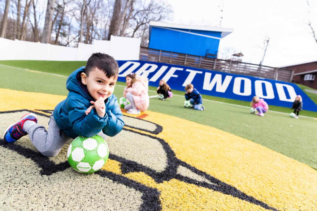 Kids on a soccer field doing arm exercises on soccer balls.